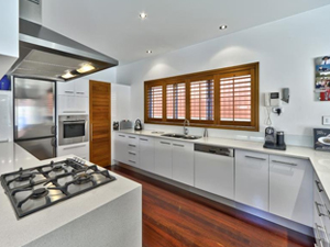 A modern kitchen featuring white cabinetry and a sleek design. The kitchen is equipped with stainless steel appliances, including an oven, microwave, and a gas cooktop on an island countertop. Large windows with wooden shutters allow natural light to fill the space, enhancing the warm wooden flooring. The overall aesthetic is contemporary and well-organised, with a focus on functionality.