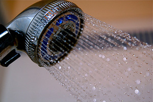 The image shows a close-up view of a showerhead with water droplets cascading from it. The showerhead features a metallic design with a circular face, and water sprays out in a fine mist, creating a refreshing atmosphere.