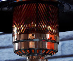 The image shows a close-up of a gas patio heater. It has a cylindrical metal base with a shiny chrome finish and a perforated heat shield that glows orange, indicating it is in use. The background appears to be a textured, possibly brick wall.