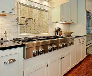 The image features a modern kitchen with sleek white cabinetry and a stainless steel gas stove. The stove has multiple burners and a polished black countertop. Behind the stove, there's a decorative mosaic backsplash, and the kitchen is well-lit with soft overhead lighting. A few bottles and a bowl of fruits are displayed on the counter, adding a warm touch to the contemporary design.