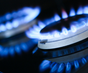The image features two gas burners ignited, exhibiting bright blue flames dancing atop a sleek, modern gas stove. The burners are silver and round, reflecting the flames in a polished surface of the stove.