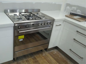 The image showcases a stainless steel gas stove with multiple burners and an oven. It is installed in a modern kitchen with white cabinetry and wooden flooring. The stove features a sleek design, with controls positioned at the front and an array of knobs for adjusting the burners.