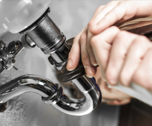 A close-up image showing a person's hands gripping a wrench while adjusting a metal pipe fitting under a sink. The focus is on the shiny chrome plumbing components and the tools being used.
